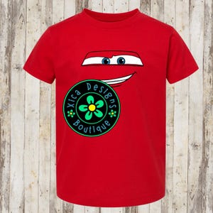 Pu&ograve; includere: Una maglietta rossa con un design di auto a cartoni animati, con occhi blu e una bocca sorridente. La maglietta presenta anche un logo circolare con il testo "Xica Designs Boutique" e un fiore verde e giallo.