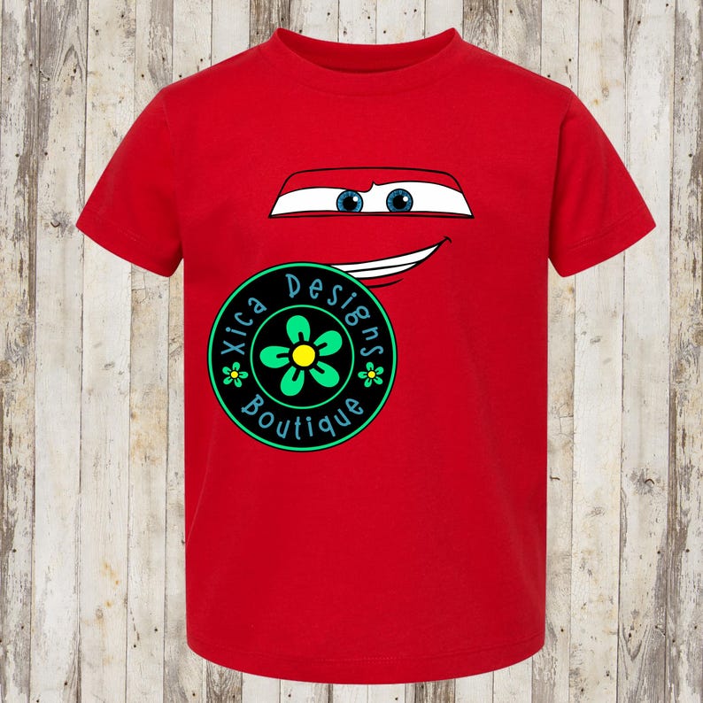 Pu&ograve; includere: Una maglietta rossa con un design di auto a cartoni animati, con occhi blu e una bocca sorridente. La maglietta presenta anche un logo circolare con il testo "Xica Designs Boutique" e un fiore verde e giallo.