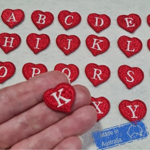 May include: Red heart-shaped embroidered patches with white capital letters. The letter "K" is held in a hand. The patches are arranged on a light-coloured surface. The text "Made in Australia" is visible.