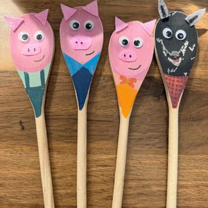 May include: Four wooden spoons decorated as characters. Three spoons are painted pink with felt ears and googly eyes, wearing painted outfits. One spoon is painted black with grey ears and googly eyes, featuring a painted red and black checkered scarf.