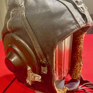 Peut inclure: Un casque d'aviateur en cuir marron avec une doublure en fourrure et un microphone attaché. Le casque a une visière transparente et une sangle avec une boucle.