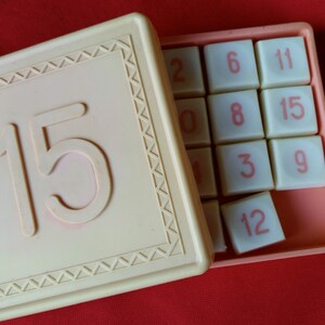 Peut inclure: Un jeu de puzzle coulissant en plastique blanc. Le puzzle comporte 15 tuiles numérotées et un espace vide. Le jeu est ouvert pour révéler les tuiles et le numéro 15 est sur le couvercle.