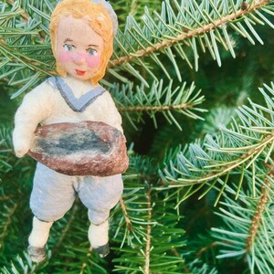 May include: A vintage Christmas tree decoration of a child figure holding a loaf of bread. The figure has a painted face, blonde hair, and is dressed in a white and blue outfit. The ornament is hanging on a green Christmas tree.