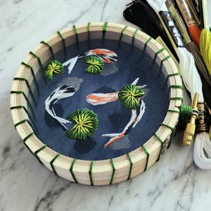 May include: Embroidered koi carp and lily pads in a round wooden hoop. The design features white, orange, and black koi carp swimming in a blue pond with green lily pads. Embroidery threads in various colours are visible.