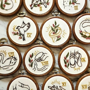 Op de afbeelding: Een verzameling geborduurde hoepels met diverse ontwerpen. Elke hoepel heeft een ander beeld, waaronder een vogel, een zwaan en balletschoenen, met gouden en rode accenten. De hoepels zijn geplaatst in houten frames met metalen sluitingen.