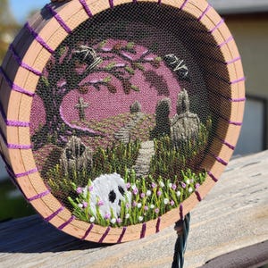 Può includere: Scena di Halloween ricamata in un telaio di legno. L'opera raffigura un cimitero con lapidi, alberi e un teschio in primo piano. Il telaio è rifinito con filo viola e ha una vite dorata.