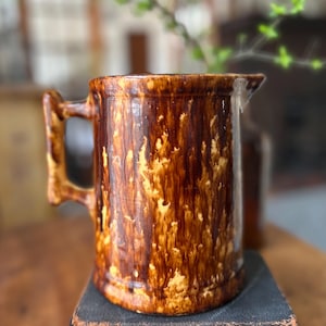 May include: A brown and yellow glazed ceramic pitcher with a handle. The pitcher has a mottled brown and yellow glaze with a rustic look. The pitcher is sitting on top of a stack of books.