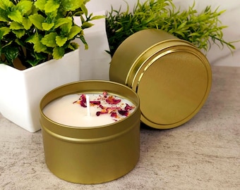 May include: A round gold metal candle tin containing white wax with a single cotton wick and scattered dried pink and red flower petals on the surface. A matching gold metal lid is positioned upright next to the open candle container.