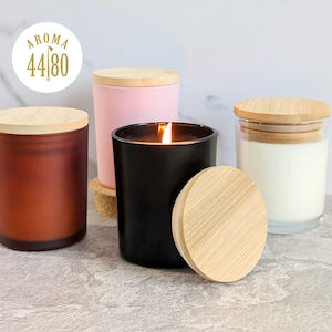 May include: Four cylindrical candles with bamboo lids. One is matte black and lit, one is matte rust-brown, one is matte pastel pink, and one is clear glass with white wax. A circular logo in the top left corner reads AROMA 44 80.