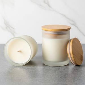 May include: Two frosted glass candles with wooden lids. One candle is upright, the other is on its side. The candles have a creamy white wax and a natural wood lid. The background is a light gray surface with a white marble pattern.