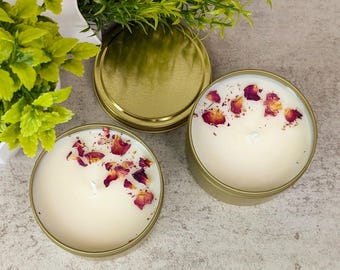 May include: Two round gold metal tins containing white wax candles with a single centered wick. The surface of each candle is decorated with scattered dried pink and red rose petals. One matching gold metal lid is placed between the two candles.