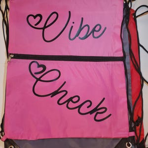 May include: Pink drawstring backpack with the words "Vibe Check" in black script. The backpack has a black zipper and black drawstrings. A red and gray backpack is partially visible behind the pink one.