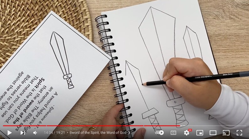 May include: A spiral-bound sketchbook featuring black-and-white line drawings of three swords, with a hand using a grey pencil to draw a fourth sword. Beside it, a printed sheet displays a sword illustration and text about the Sword of the Spirit.