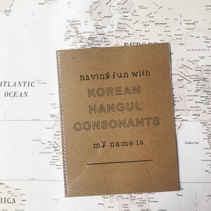 May include: A brown notebook with a spiral binding. The cover has the text "having fun with KOREAN HANGUL CONSONANTS my name is" printed on it. The notebook is sitting on a world map.