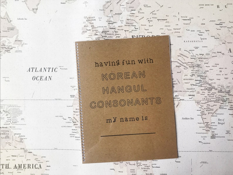 May include: A brown notebook with a spiral binding. The cover has the text "having fun with KOREAN HANGUL CONSONANTS my name is" printed on it. The notebook is sitting on a world map.