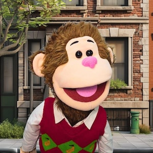 May include: A plush monkey puppet with brown fur, a pink nose, and a wide smile. It wears a red sweater vest with a green and yellow banana pattern over a white collared shirt. The background shows a brick building and a tree.