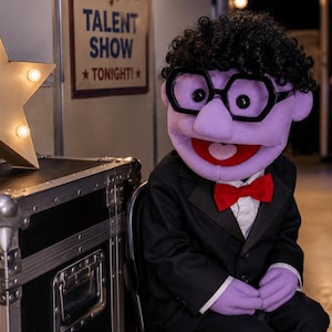 Op de afbeelding: Een paarse handpop met krullend zwart haar en een zwarte zeshoekige bril, gekleed in een zwart colbert, wit overhemd en een rode vlinderdas. De pop staat bij een bord met de tekst TALENT SHOW TONIGHT en een verlichte ster als rekwisiet.