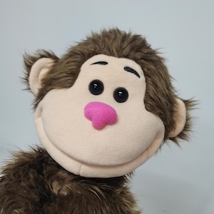 May include: A plush monkey puppet with a light tan face, brown fur, and a pink nose. The monkey has large black eyes and a smiling expression. The puppet is designed to be held and manipulated.