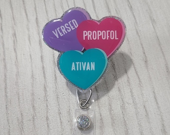 May include: A silver glitter badge reel with three heart-shaped charms. The charms are purple, pink, and teal. The purple charm says "VERSED", the pink charm says "PROPOFOL", and the teal charm says "ATIVAN".