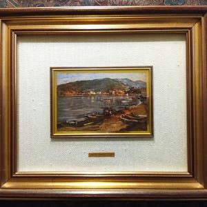 Op de afbeelding: Een ingelijst landschapsschilderij met een kustlandschap met boten op de oever en een stad in de verte. Het schilderij is gevat in een goudkleurige lijst, gemonteerd op een getextureerde witte achtergrond en ingesloten in een grotere, sierlijke gouden lijst.