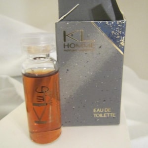 May include: A small glass vial of amber-colored liquid with the text "Gim" and "KL Homme" perfume packaging. The box is gray with gold accents and the words "EAU DE TOILETTE".