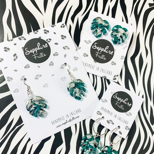 May include: Three pairs of earrings featuring a green and silver monstera leaf design. The earrings are handmade in England and are available in stud, hoop, and dangle styles.