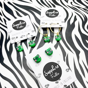 May include: A set of four green frog earrings with pink accents. The earrings are handmade in England and come on a card with the text "Sapphire Frills" and "Handmade in England".