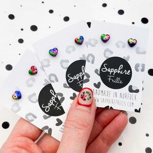 May include: Three sets of heart-shaped stud earrings with a colorful, iridescent finish. Each set is displayed on a white card with a black and white leopard print design and the text "Sapphire Frills".