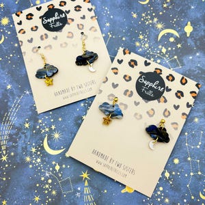 Pu&ograve; includere: Due paia di orecchini a forma di nuvola su cartoncini espositivi. Ogni orecchino presenta una nuvola marmorizzata blu scuro e nera con una piccola stella glitterata dorata o un ciondolo a mezzaluna bianca, appesi a componenti in metallo dorato.