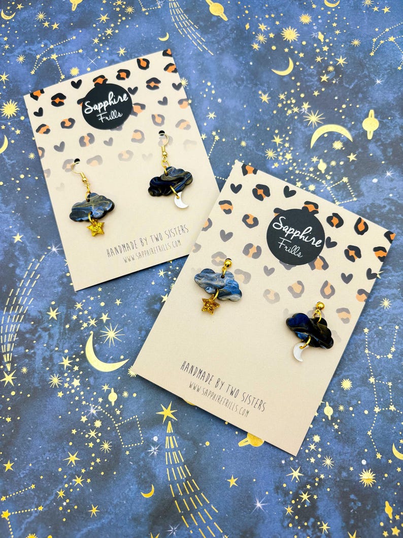 Pu&ograve; includere: Due paia di orecchini a forma di nuvola su cartoncini espositivi. Ogni orecchino presenta una nuvola marmorizzata blu scuro e nera con una piccola stella glitterata dorata o un ciondolo a mezzaluna bianca, appesi a componenti in metallo dorato.