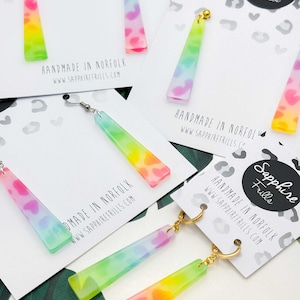 May include: Multiple pairs of dangle earrings featuring long, rectangular, translucent acrylic pendants with a multi-coloured leopard print pattern in neon pink, yellow, green, and blue, attached to gold-tone metal hooks on branded display cards.