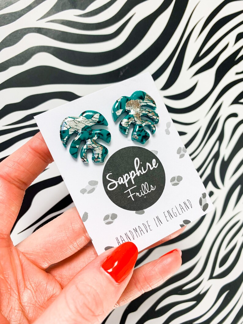 May include: A pair of teal green and silver monstera leaf earrings. The earrings are handmade in England by Sapphire Frills.