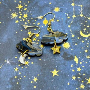 Pu&ograve; includere: Un paio di orecchini pendenti con nuvole marmorizzate blu scuro e nere. Un orecchino ha un ciondolo a forma di mezzaluna bianca, l'altro una stella glitterata dorata. Entrambi sono appesi a ganci per orecchini in metallo color oro.