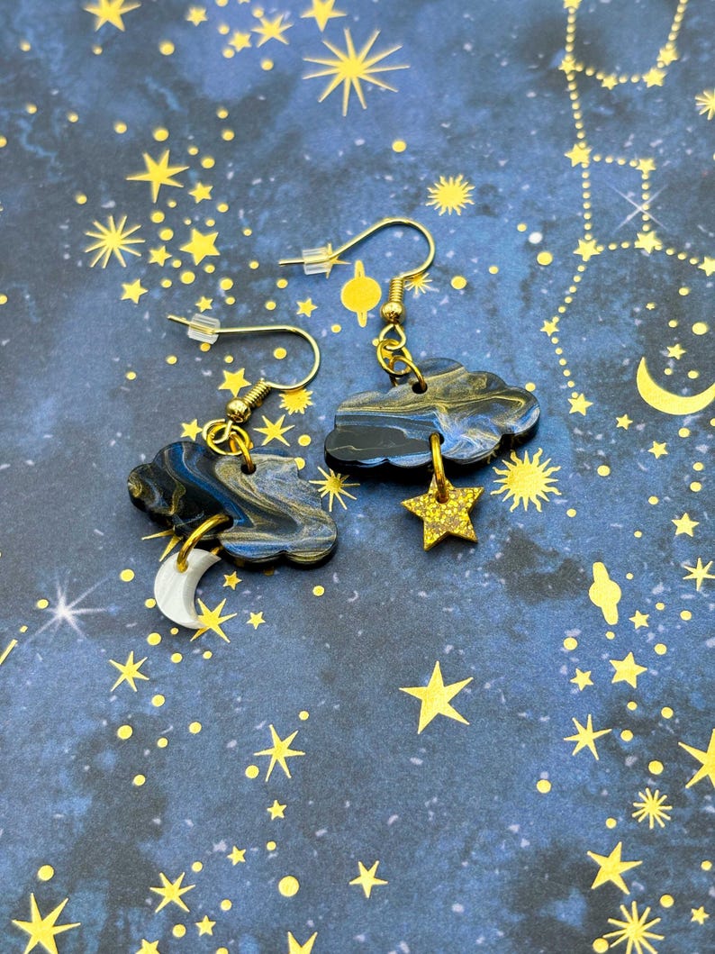 Pu&ograve; includere: Un paio di orecchini pendenti con nuvole marmorizzate blu scuro e nere. Un orecchino ha un ciondolo a forma di mezzaluna bianca, l'altro una stella glitterata dorata. Entrambi sono appesi a ganci per orecchini in metallo color oro.