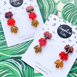 Peut inclure: Une paire de boucles d'oreilles avec trois feuilles d'érable superposées dans des tons de rouge, orange et or. Les boucles d'oreilles sont faites à la main en Angleterre et sont présentées sur une carte blanche avec le nom de la marque "Sapphire Frills" et l'adresse du site Web.