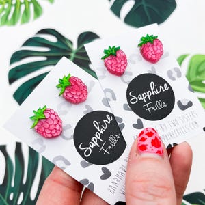 May include: A pair of pink raspberry earrings with green leaves. The earrings are handmade by Sapphire Frills, a company owned by two sisters. The earrings are displayed on a white card with a black circle that says "Sapphire Frills".