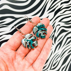 May include: A pair of silver and teal green monstera leaf earrings. The leaves have a silver crackle finish.