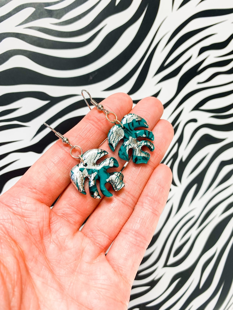 May include: A pair of silver and teal green monstera leaf earrings. The leaves have a silver crackle finish.