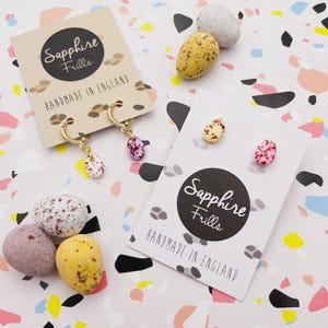 May include: Gold hoop earrings and stud earrings with speckled egg-shaped charms. The earrings are displayed on cards that read "Sapphire Frills Handmade in England." Several speckled egg-shaped candies in various colours are scattered around the earrings.