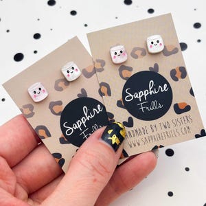 May include: Two pairs of stud earrings shaped like white marshmallows with smiling faces, pink cheeks, and black eyes. The earrings are attached to tan display cards featuring a brown leopard print pattern and the text Sapphire Frills.