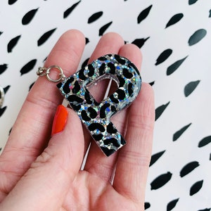 Può includere: Un portachiavi argentato con un ciondolo a forma di lettera P con stampa leopardata nera e argento.