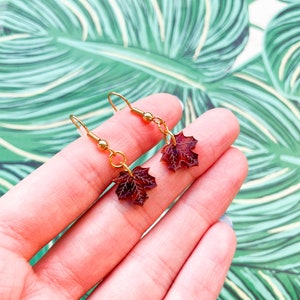 Peut inclure: Une paire de boucles d'oreilles pendantes dorées avec des breloques en forme de feuille d'érable rouge. Les boucles d'oreilles sont sur une main avec un fond vert.