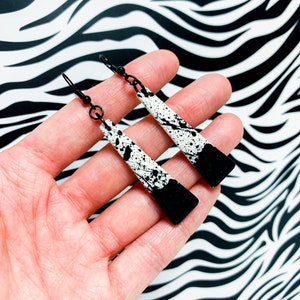 May include: A pair of black and white dangle earrings with a splatter paint design. The earrings are long and narrow, with a black metal hook.