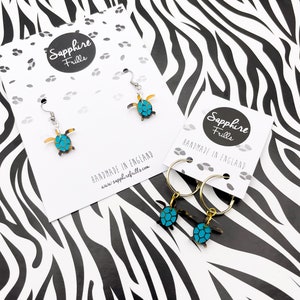 May include: Two pairs of earrings featuring a turquoise and gold turtle design. One pair is a dangle earring with a silver hook, the other is a hoop earring with a gold hoop. Both pairs are handmade in England by Sapphire Frills.