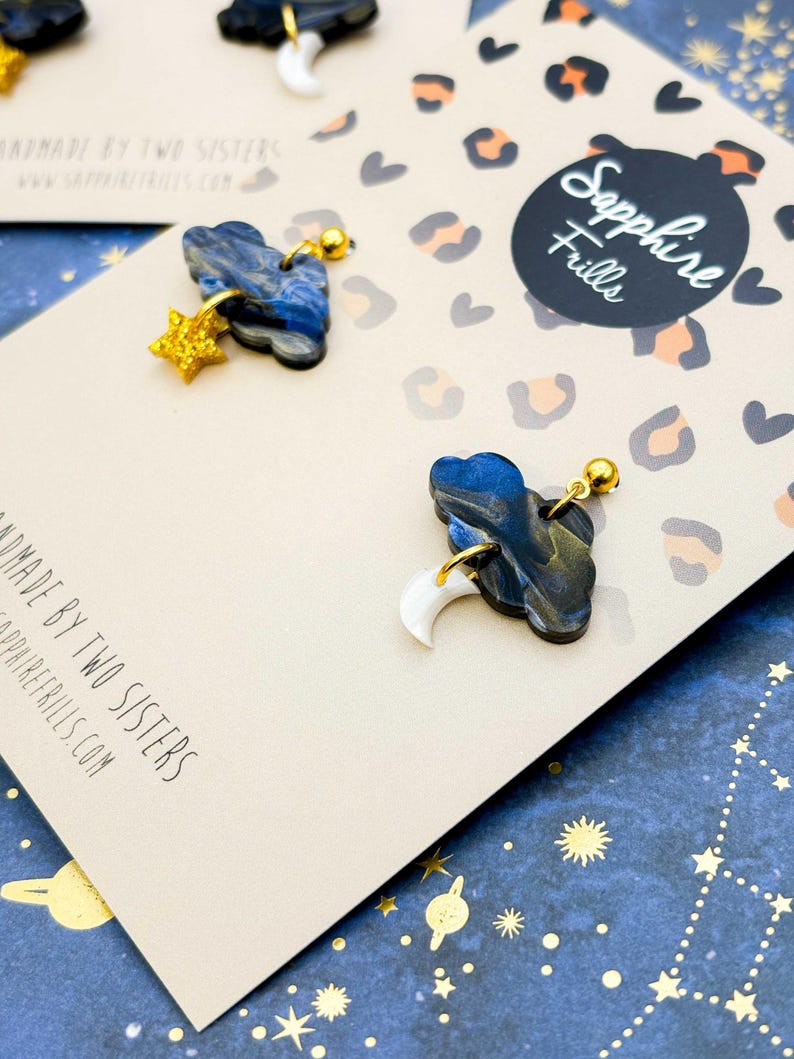 Pu&ograve; includere: Due paia di orecchini pendenti su un cartoncino beige con stampa leopardata. Ogni orecchino presenta una forma di nuvola marmorizzata blu e nera con dettagli dorati. Una nuvola ha una stella glitterata dorata, l'altra una mezzaluna bianca.