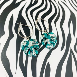 May include: A pair of silver hoop earrings with teal green monstera leaf charms. The leaves have a silver foil accent.