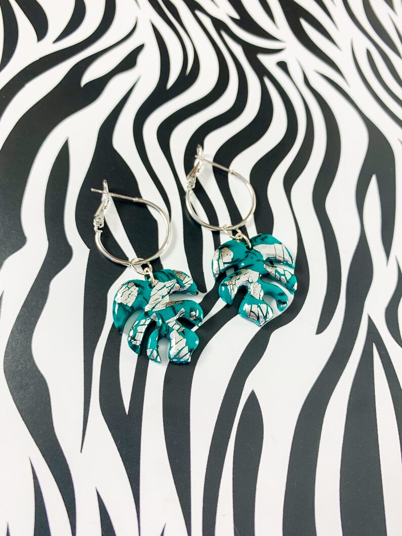 May include: A pair of silver hoop earrings with teal green monstera leaf charms. The leaves have a silver foil accent.