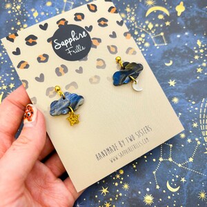 Pu&ograve; includere: Un paio di orecchini su un cartoncino con stampa leopardata che riporta il testo Sapphire Frills e Handmade by two sisters www.sapphirefrills.com. Gli orecchini hanno la forma di nuvole marmorizzate blu scuro e grigie con una stella oro e una luna bianca.