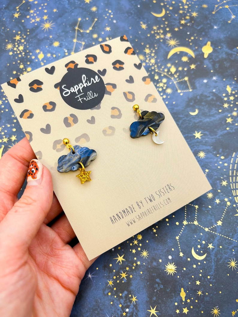 Pu&ograve; includere: Un paio di orecchini su un cartoncino con stampa leopardata che riporta il testo Sapphire Frills e Handmade by two sisters www.sapphirefrills.com. Gli orecchini hanno la forma di nuvole marmorizzate blu scuro e grigie con una stella oro e una luna bianca.