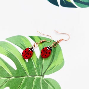May include: Ladybird earrings with red bodies and black spots, suspended from copper-coloured hooks. The earrings are displayed against a backdrop of a large green leaf pattern on a white surface.